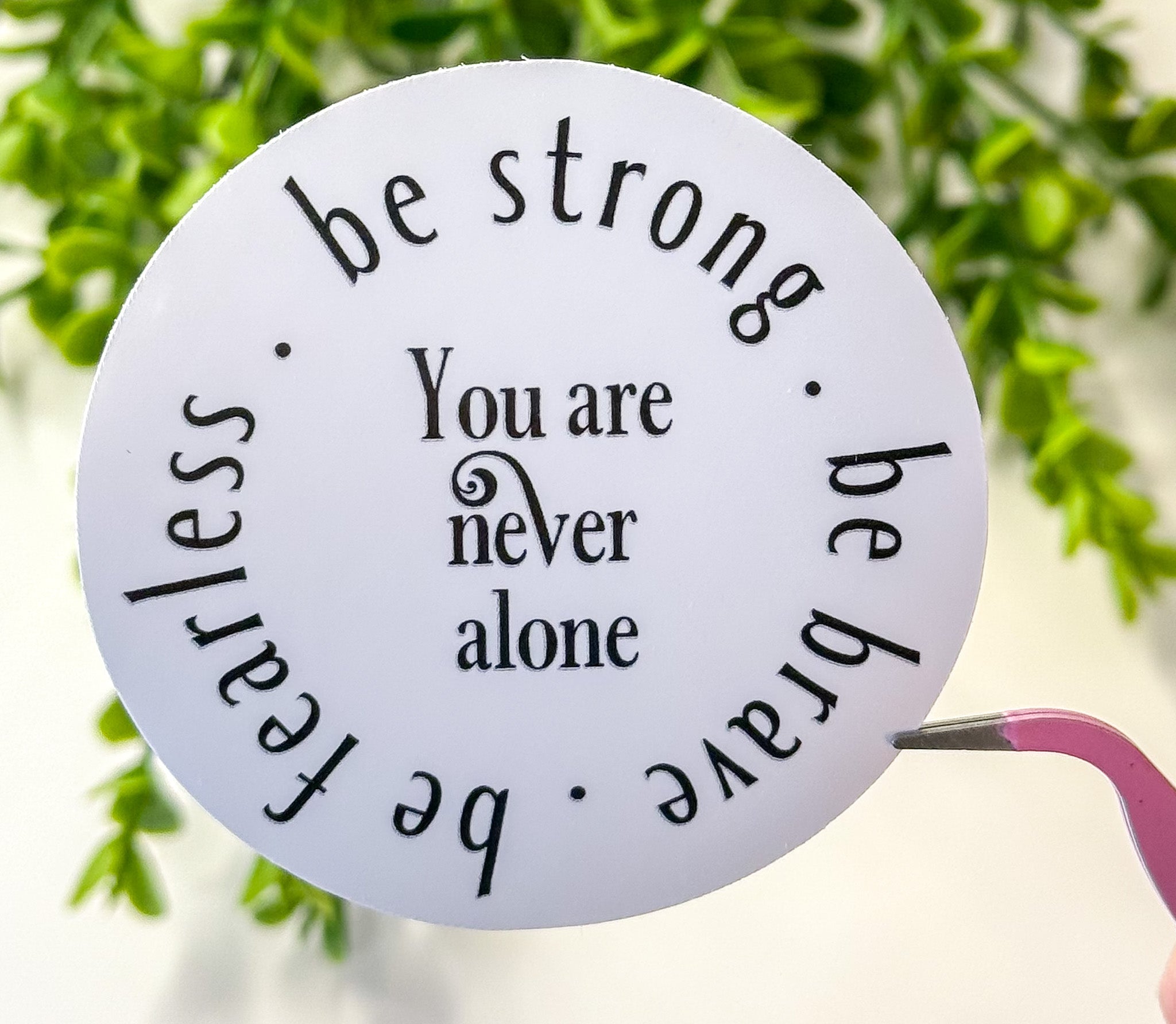 Round motivational sticker reading “be strong” and “You are never alone” in black on white