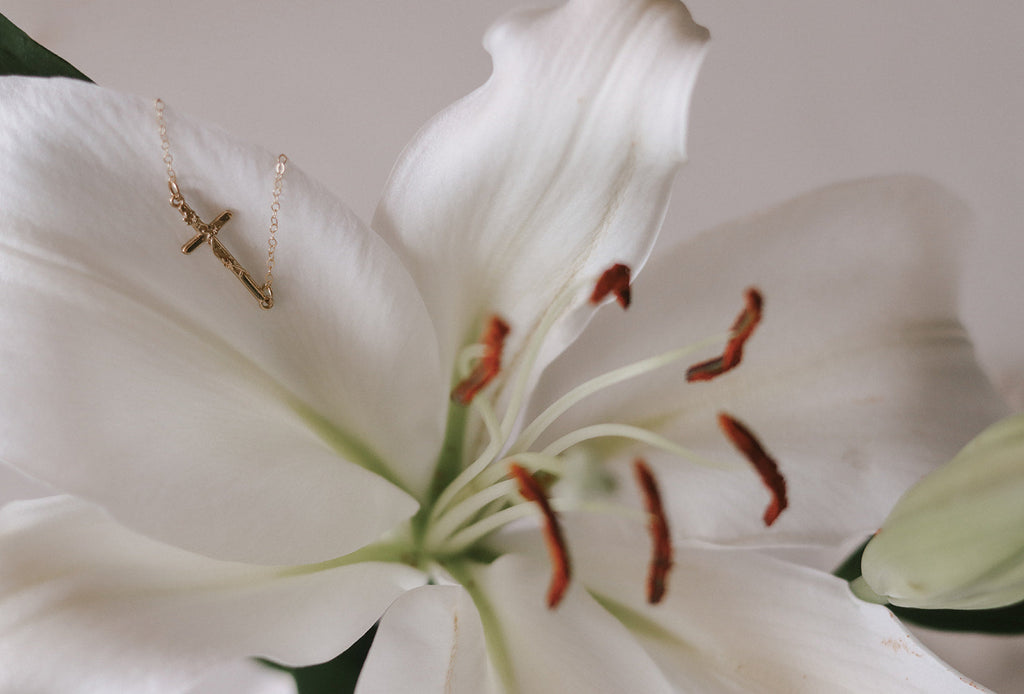 Gold cross charm bracelet draped near white lily blossoms