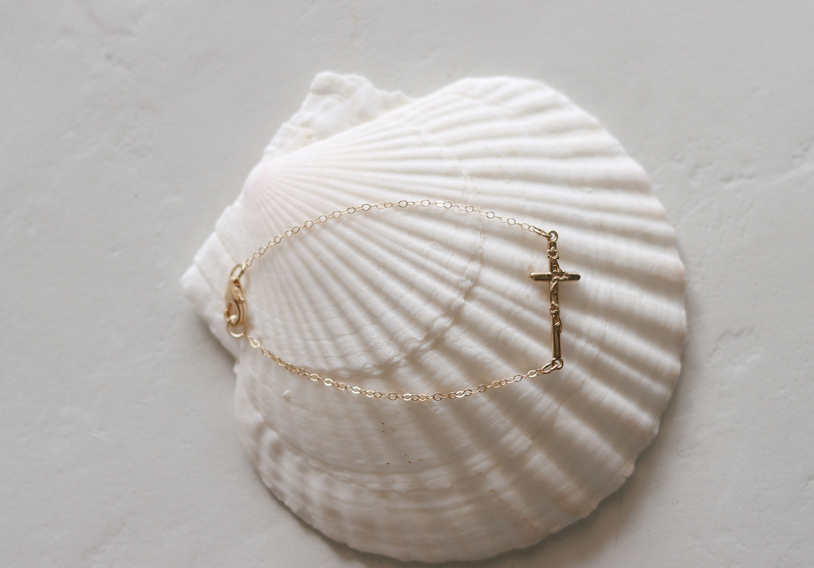 Gold-tone chain bracelet with small cross charm, resting on a white seashell