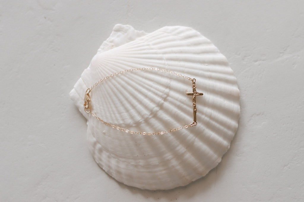 Gold-tone chain bracelet with cross charm, shown on a white seashell