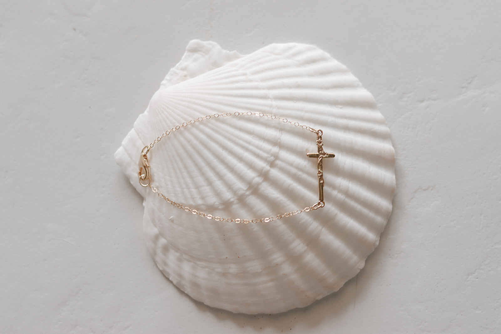 Gold-tone chain bracelet with cross charm, shown on a white seashell