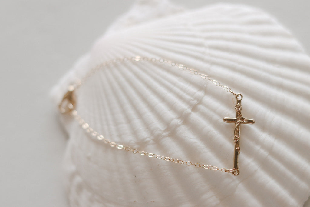 Gold-tone chain bracelet with small cross charm on a white seashell