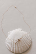 Gold-tone cross pendant necklace with fine chain, resting on a white shell dish.