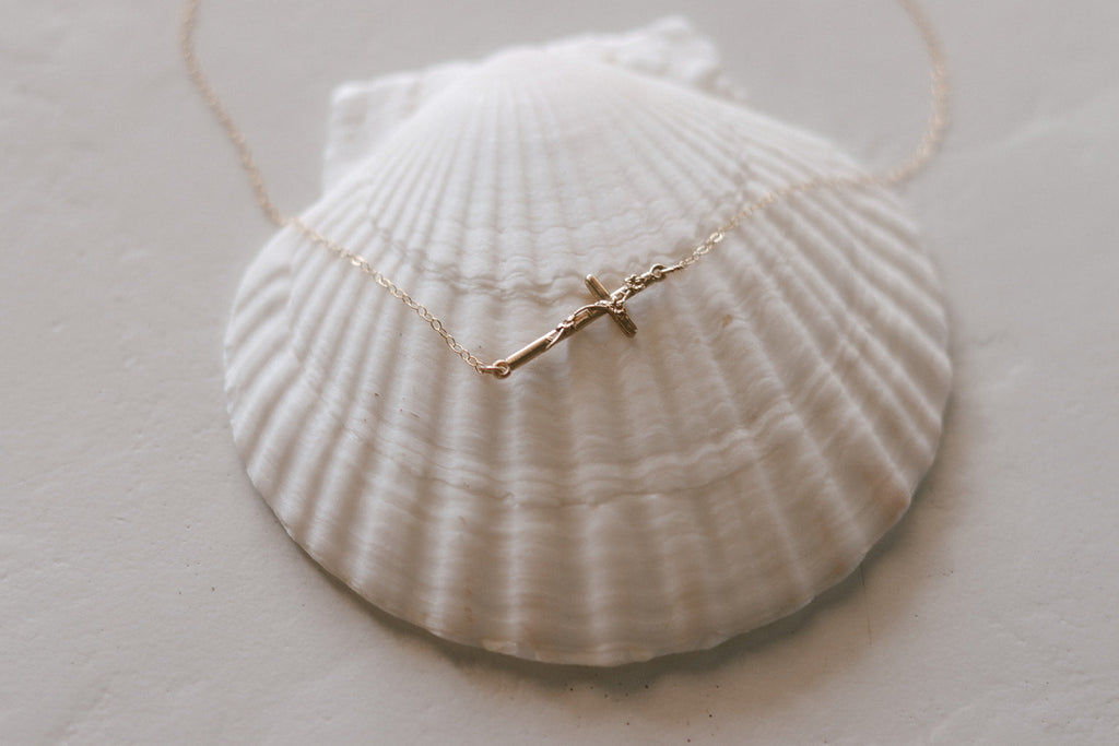 Gold-tone cross pendant necklace with fine chain on a white shell dish, pendant centered.