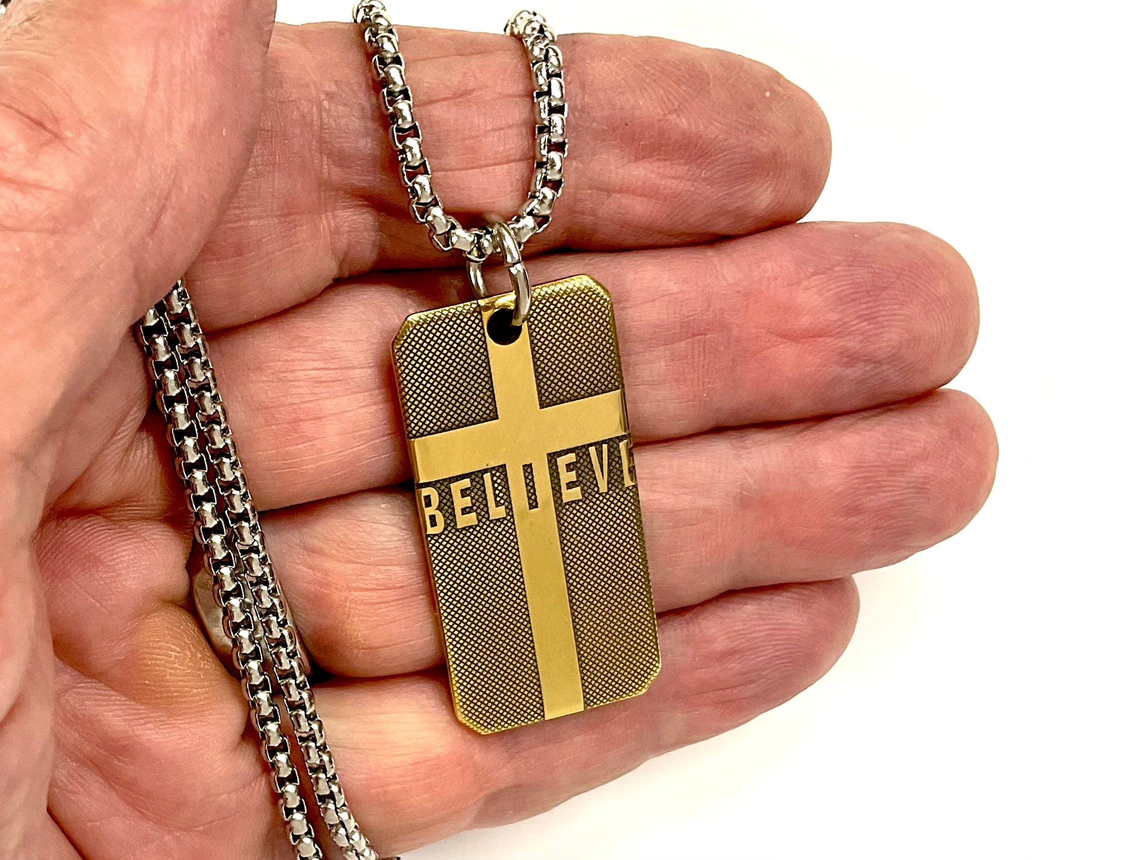 Gold-tone dog tag cross pendant with 'BELIEVE' text hanging on chain in hand