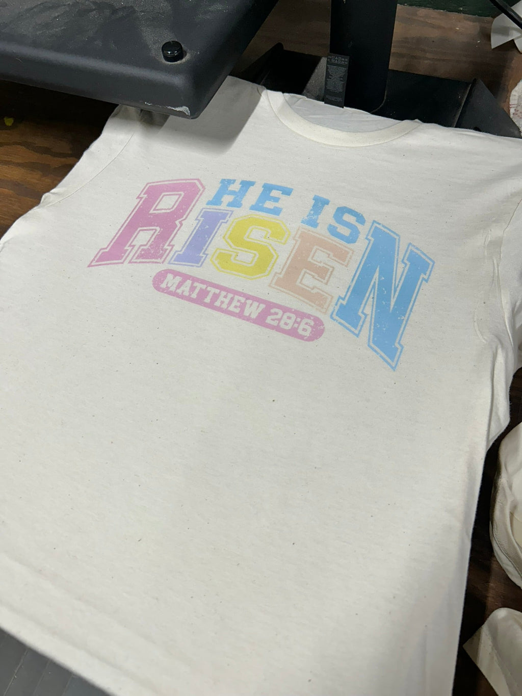 White crewneck T-shirt with pastel “He is Risen” and “Matthew 28:6” print