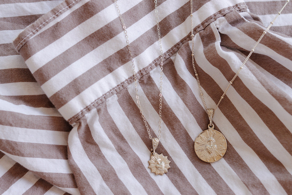 Small silver-tone and gold-tone pendant necklaces laid on striped fabric.