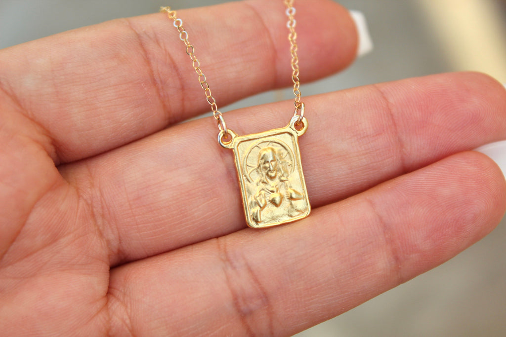 Gold-tone scapular pendant necklace with square framed medallion on fine chain, hand-held.