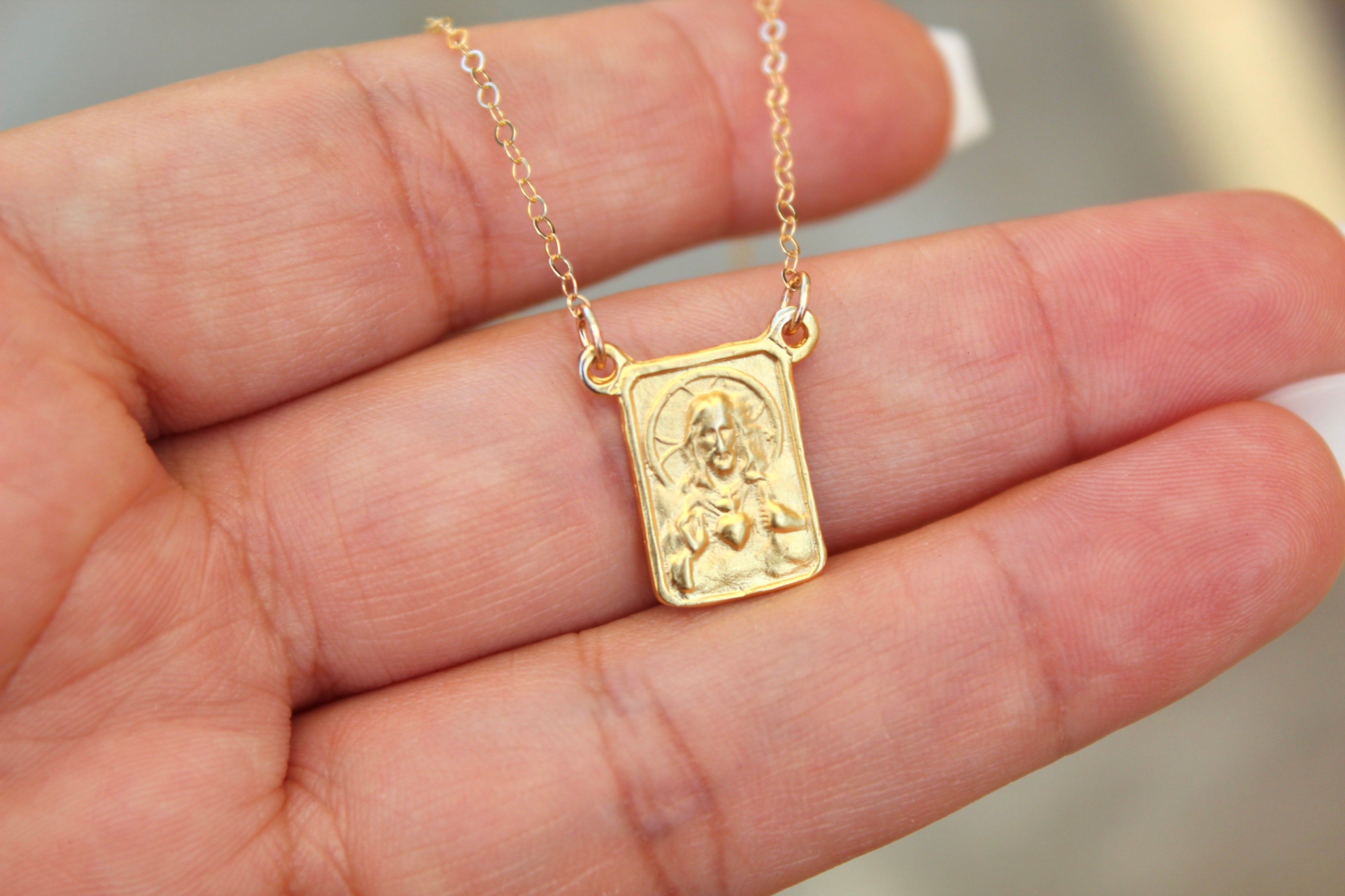 Gold-tone scapular pendant necklace with square framed medallion on fine chain, hand-held.