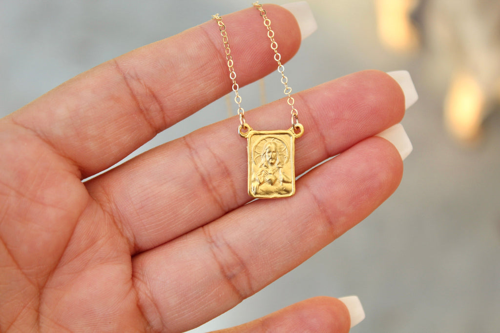 Gold-tone scapular pendant necklace with small square medallion on fine chain, held in hand.