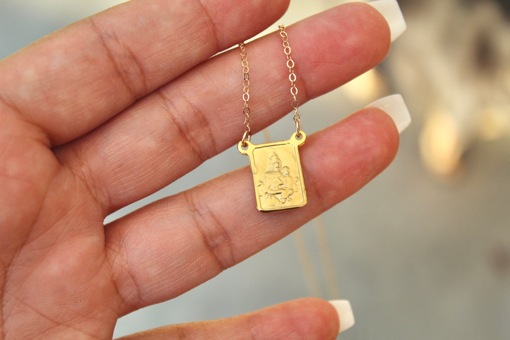 Gold-tone scapular pendant necklace with square charm on delicate chain links, hand-held.