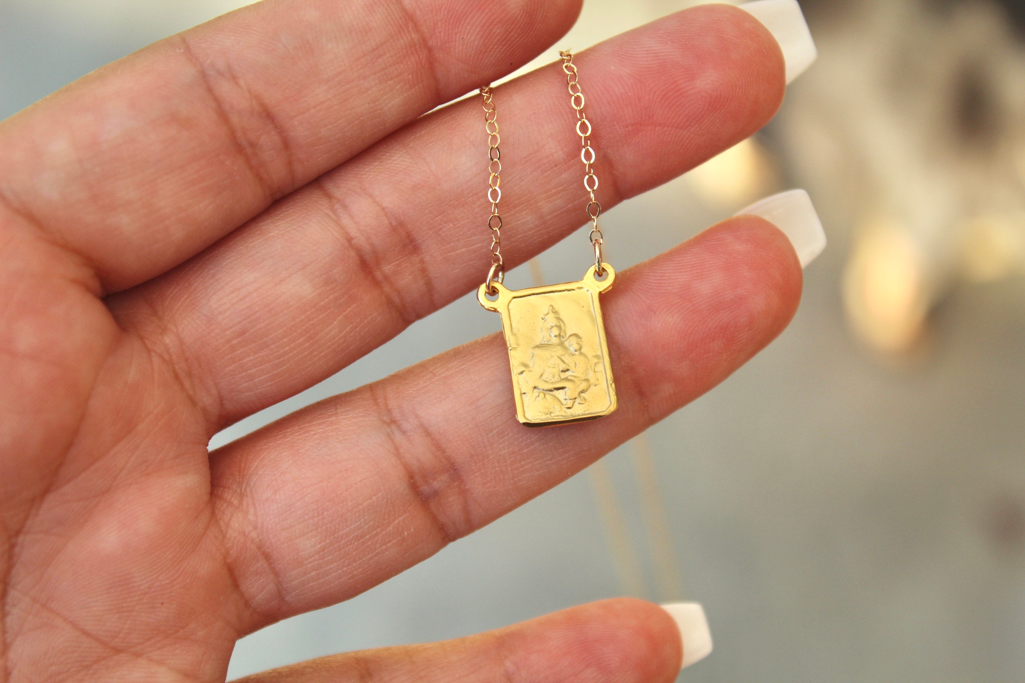 Gold-tone scapular pendant necklace with square charm on delicate chain links, hand-held.