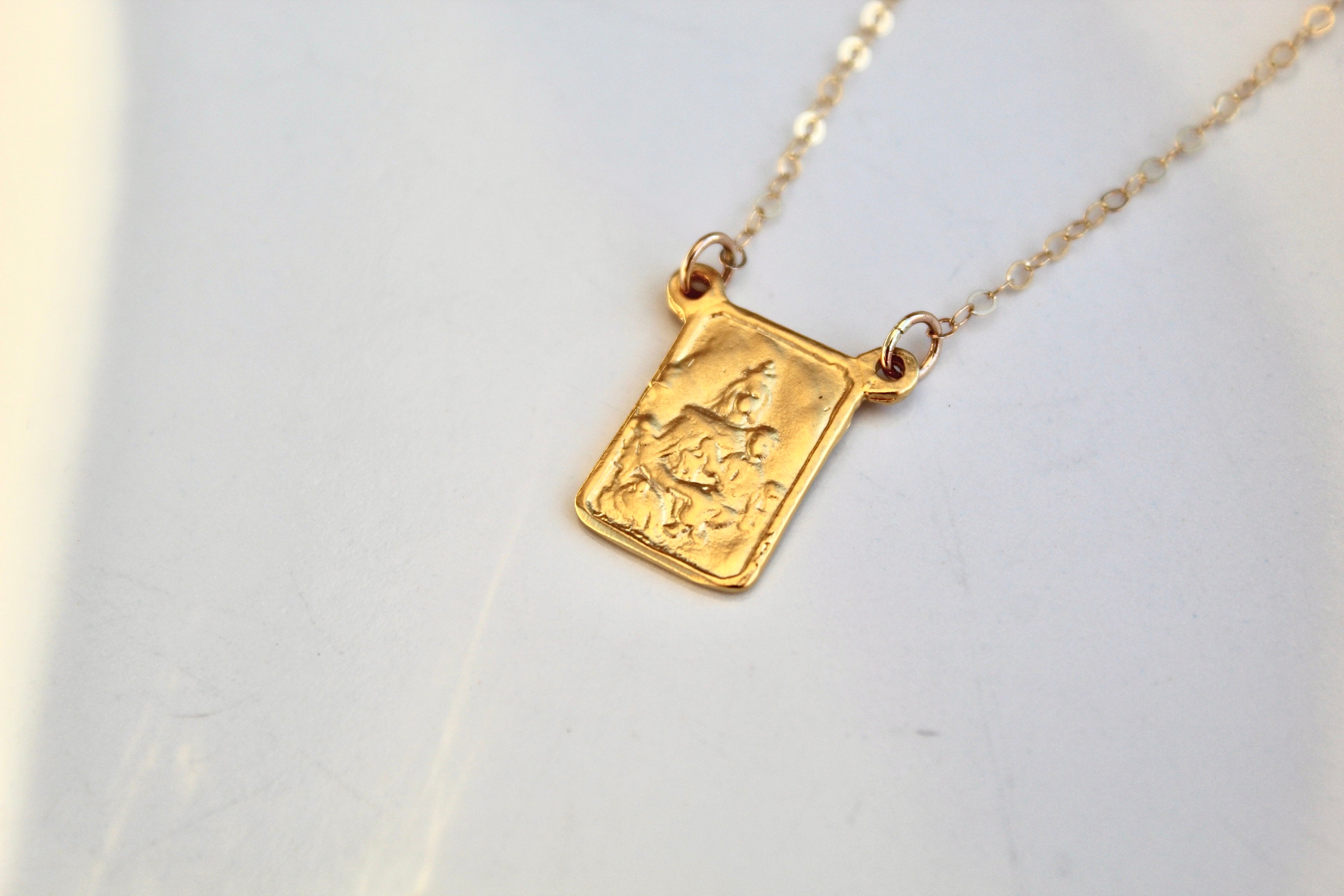 Gold-tone scapular necklace with square pendant charm and fine chain, laid flat.