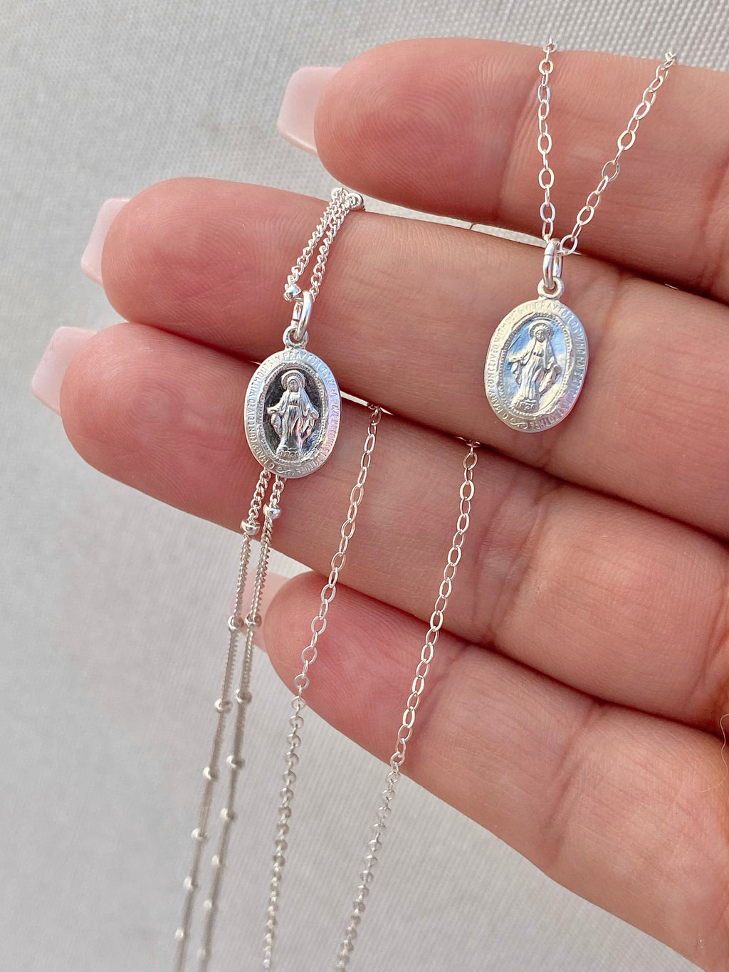 Two silver miraculous medal necklaces with oval pendants and fine linked chains, close-up