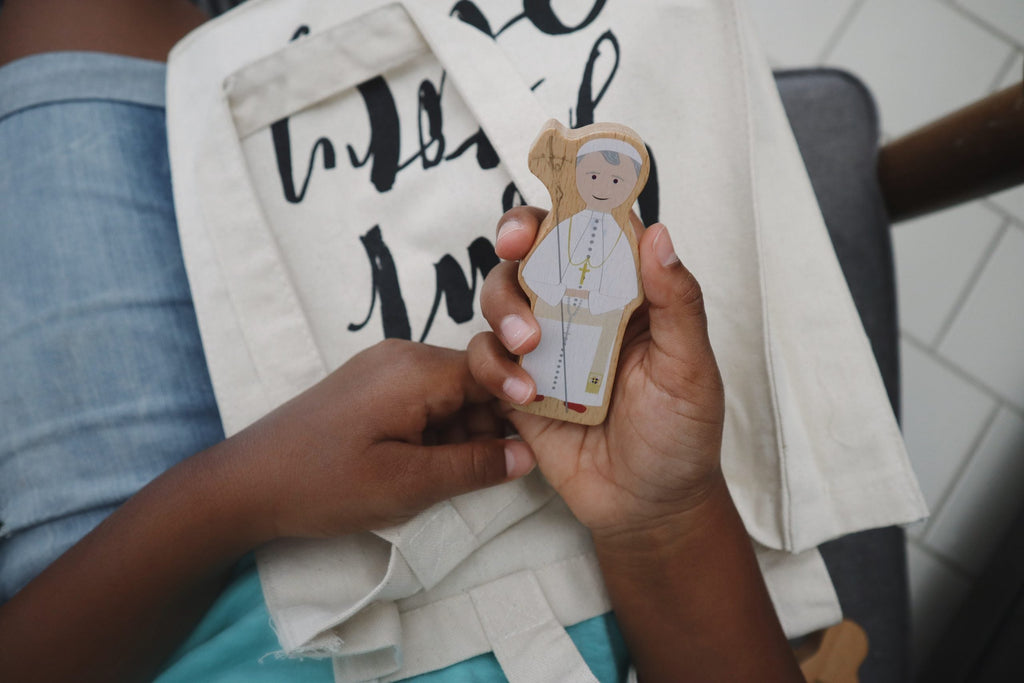 Wooden saint figure cutout held in palm above a white tote bag