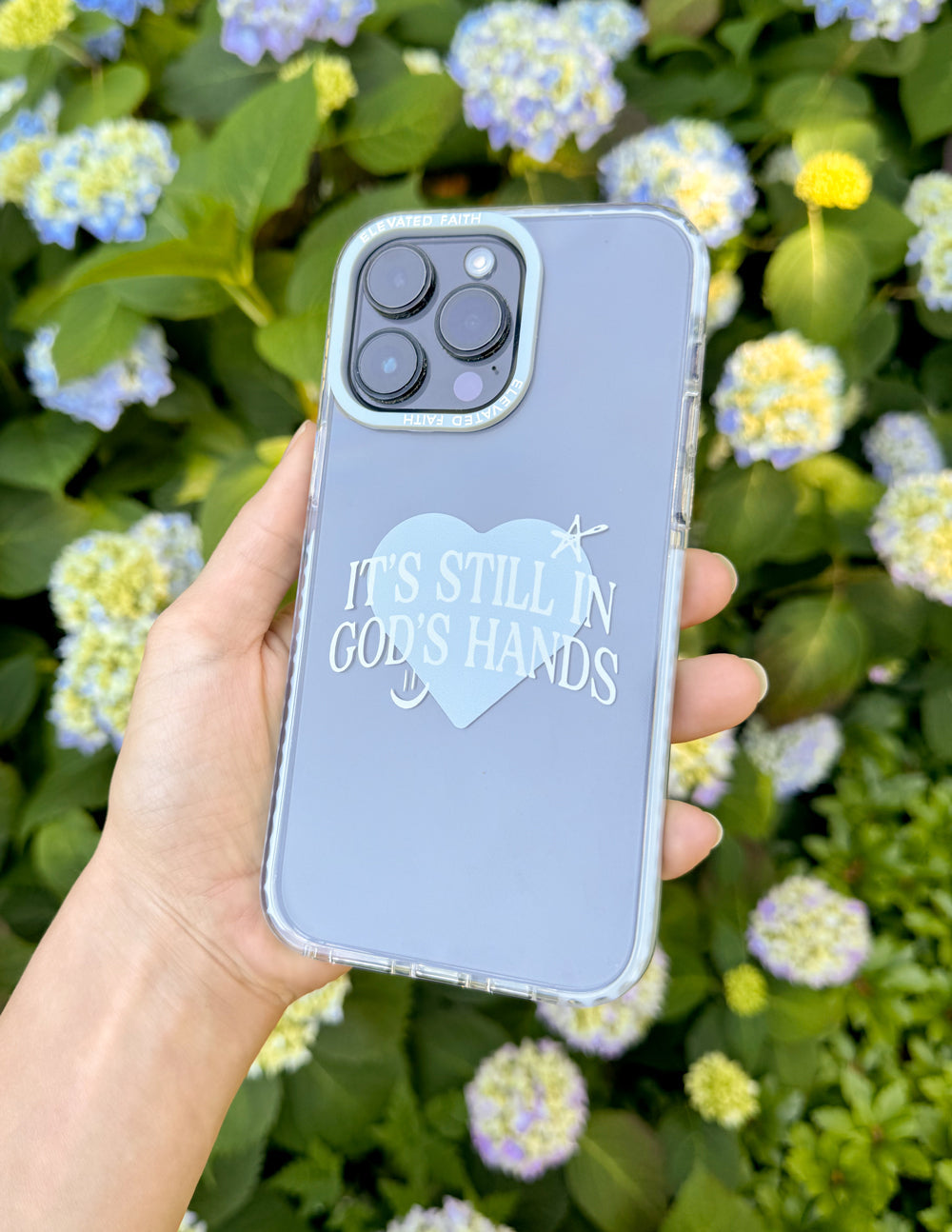 Phone case with pale blue back, heart graphic and 'It's still in God's hands' lettering