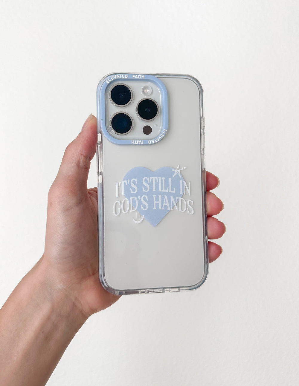 Phone case held in hand, clear light back with heart graphic and 'It's still in God's hands'