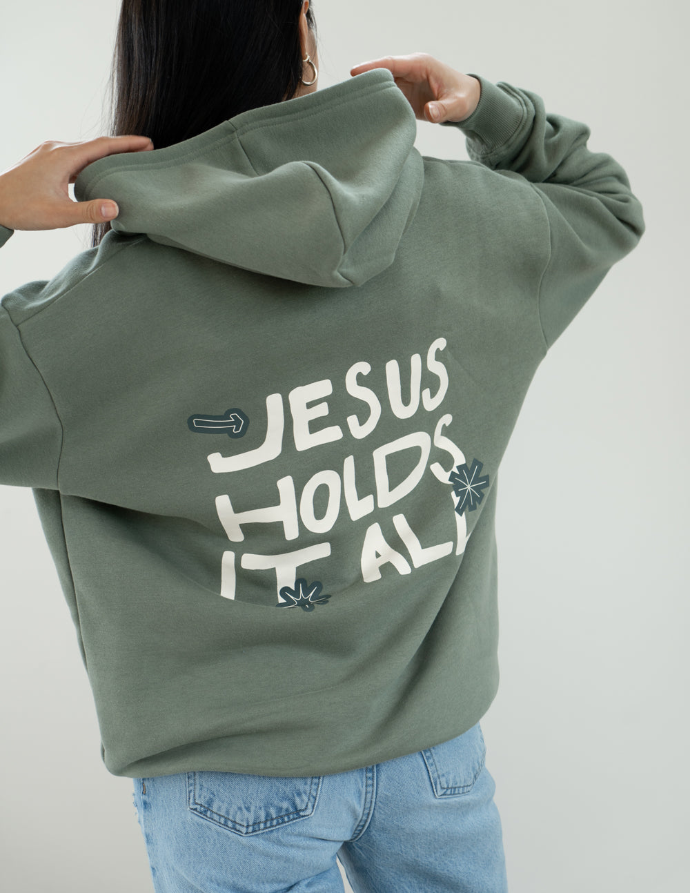Sage green hoodie with “JESUS HOLDS IT ALL” back print and hood
