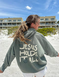 Sage green unisex hoodie with “JESUS HOLDS IT ALL” graphic on the back