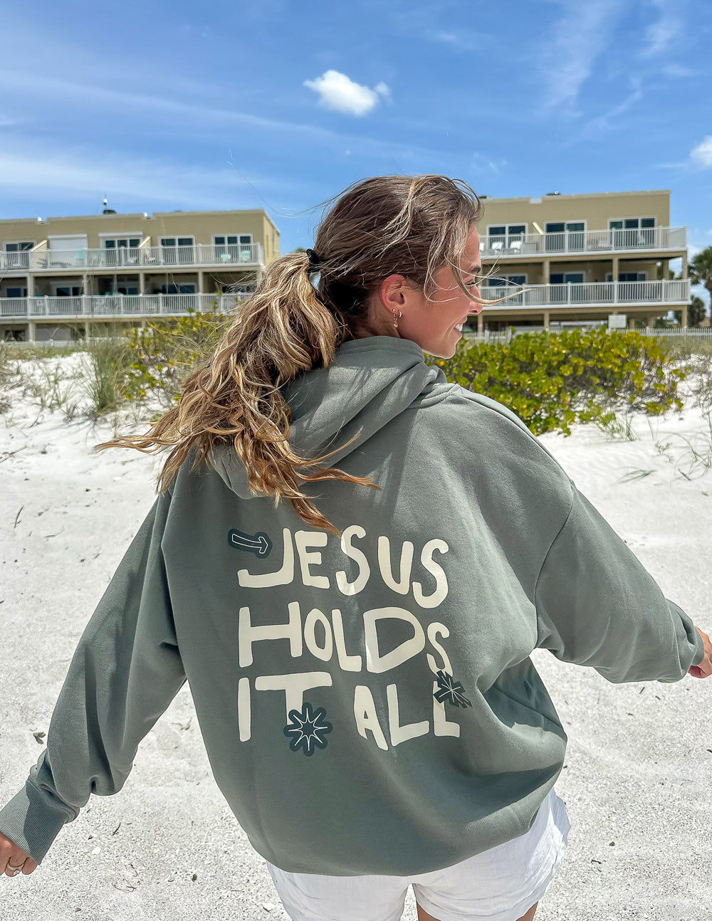 Sage green unisex hoodie with “JESUS HOLDS IT ALL” graphic on the back