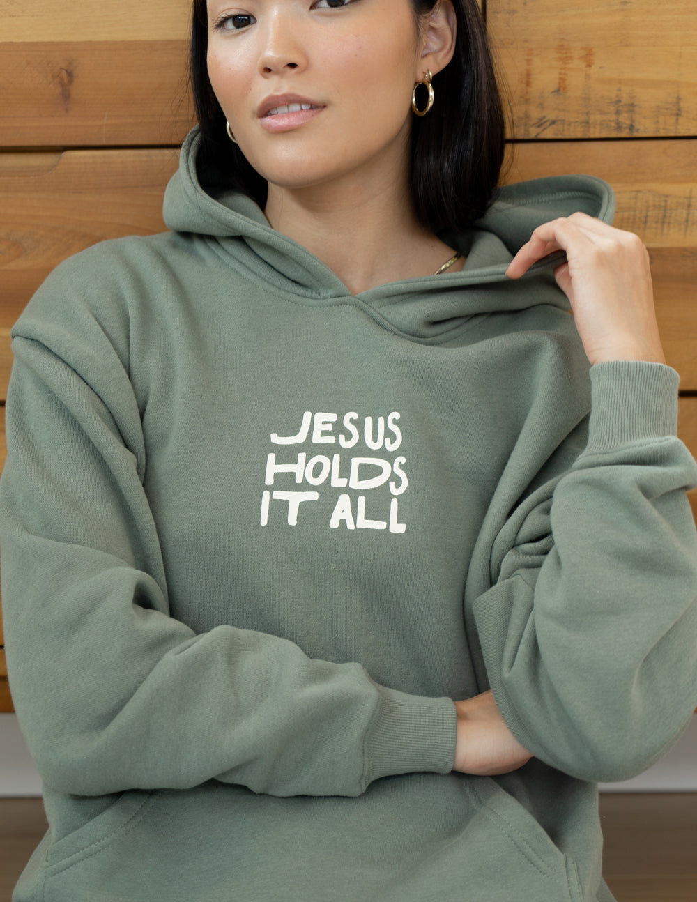 Sage green unisex hoodie with “JESUS HOLDS IT ALL” chest print