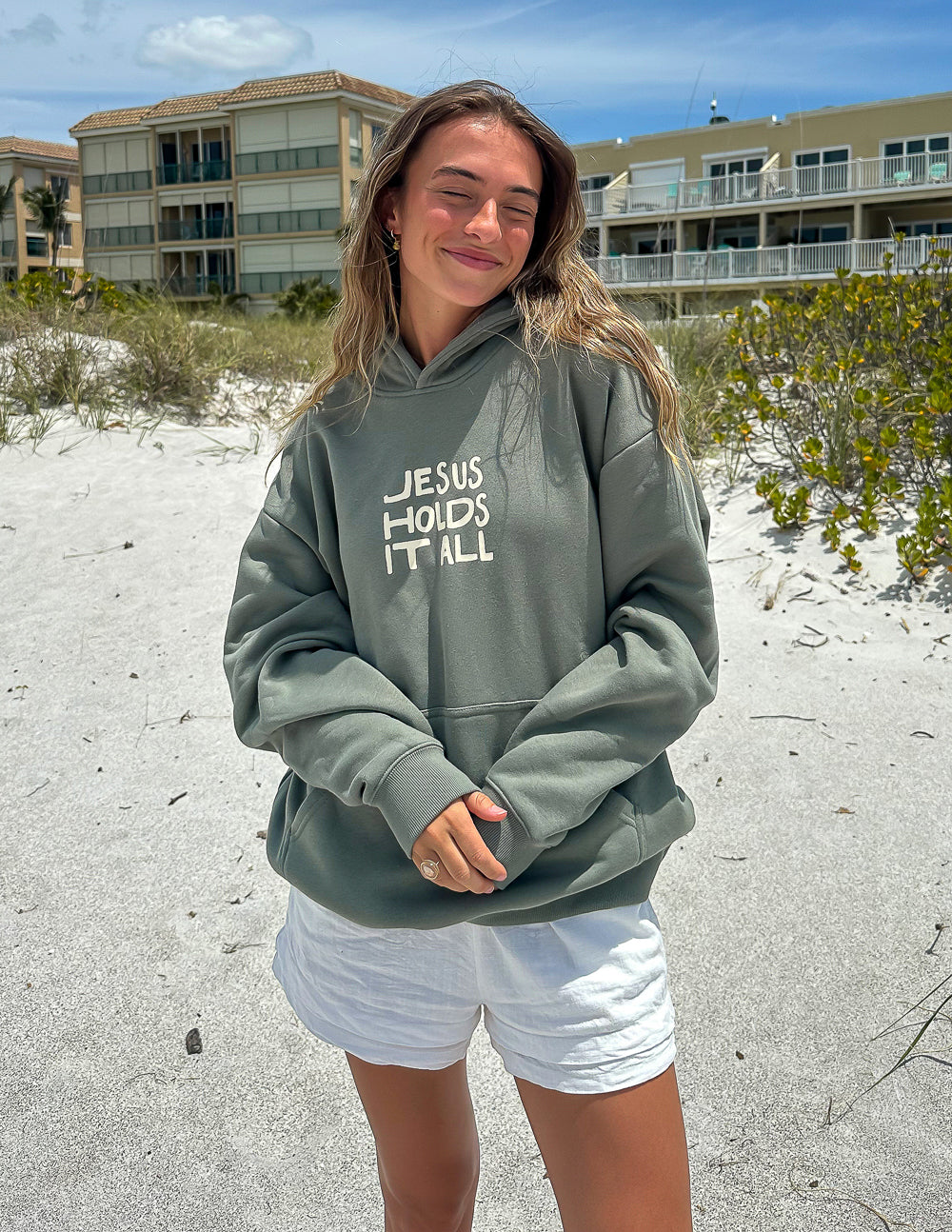 Sage green unisex hoodie with “JESUS HOLDS IT ALL” text graphic on the chest