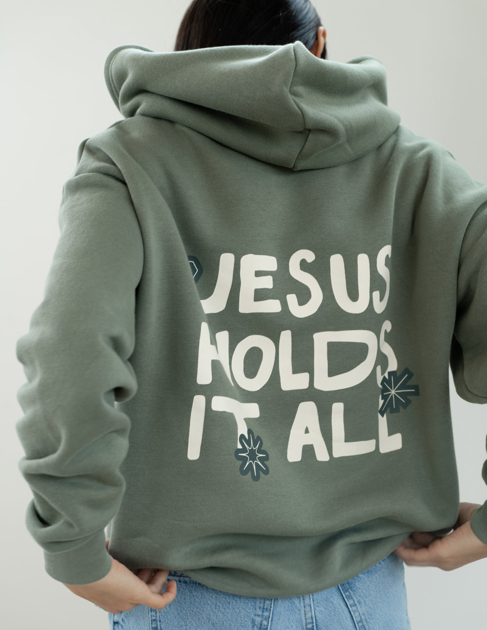Sage green unisex hoodie with “JESUS HOLDS IT ALL” graphic on the back