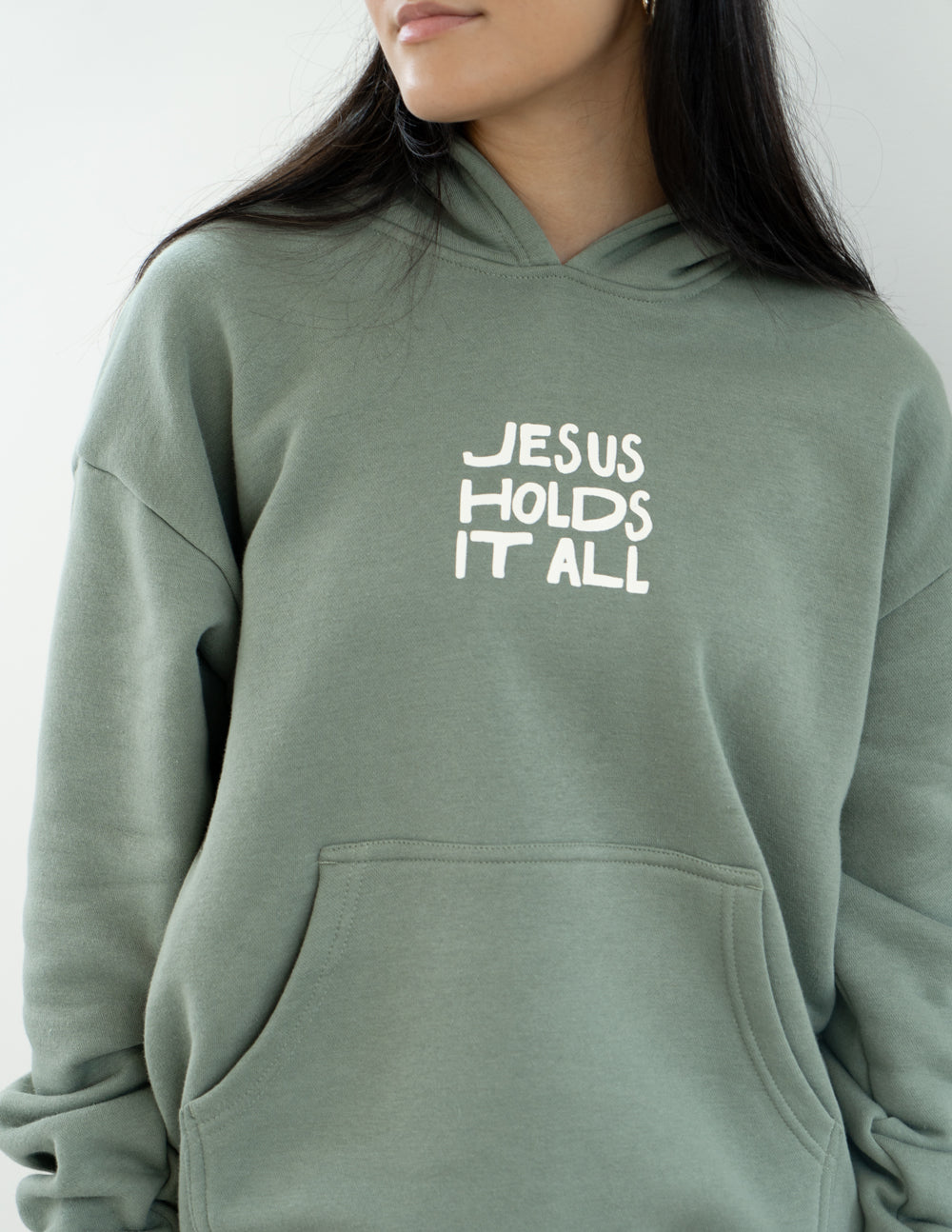 Sage green unisex hoodie with “JESUS HOLDS IT ALL” chest print
