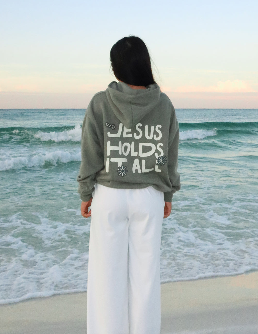 Sage green unisex hoodie with “JESUS HOLDS IT ALL” back graphic