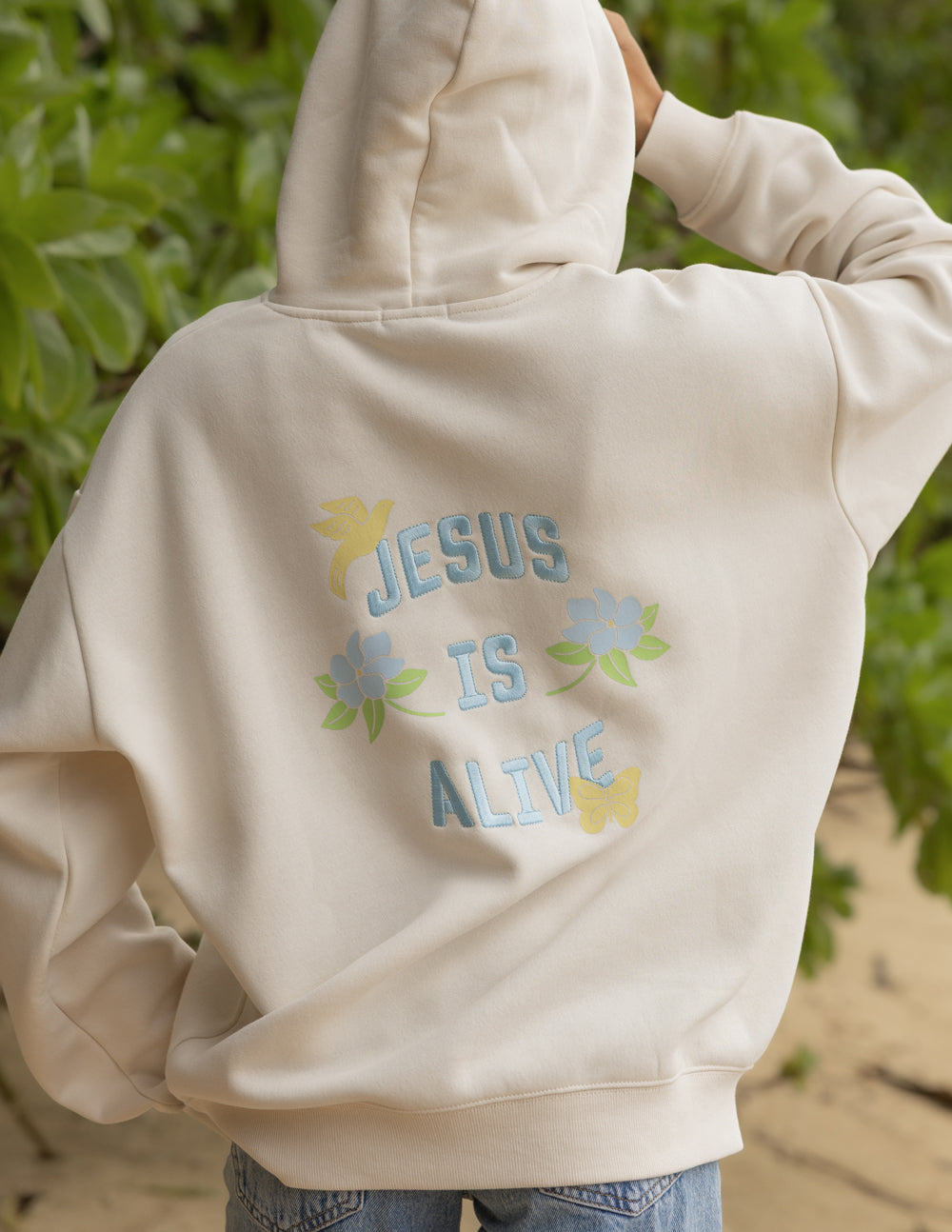 Unisex hoodie, cream, back graphic shows “JESUS IS ALIVE” with colorful flowers