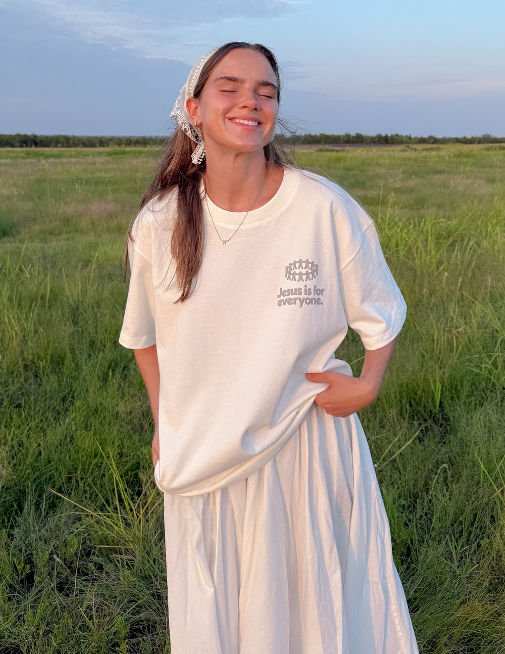 White heavyweight graphic T-shirt on woman in outdoor field, front chest logo visible