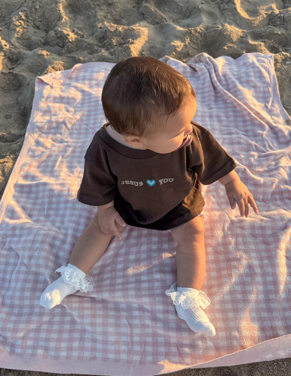 Kids brown “JESUS YOU” graphic T-shirt seen from back while lying on a blanket