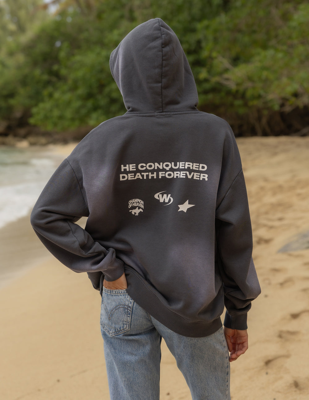 Dark gray unisex hoodie with hood and front pocket, back text “THE CONQUERED DEATH FOREVER”