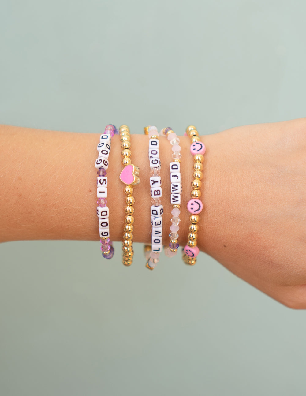 Stacked beaded bracelets on wrist with pink letter beads, gold accents, and heart charms.