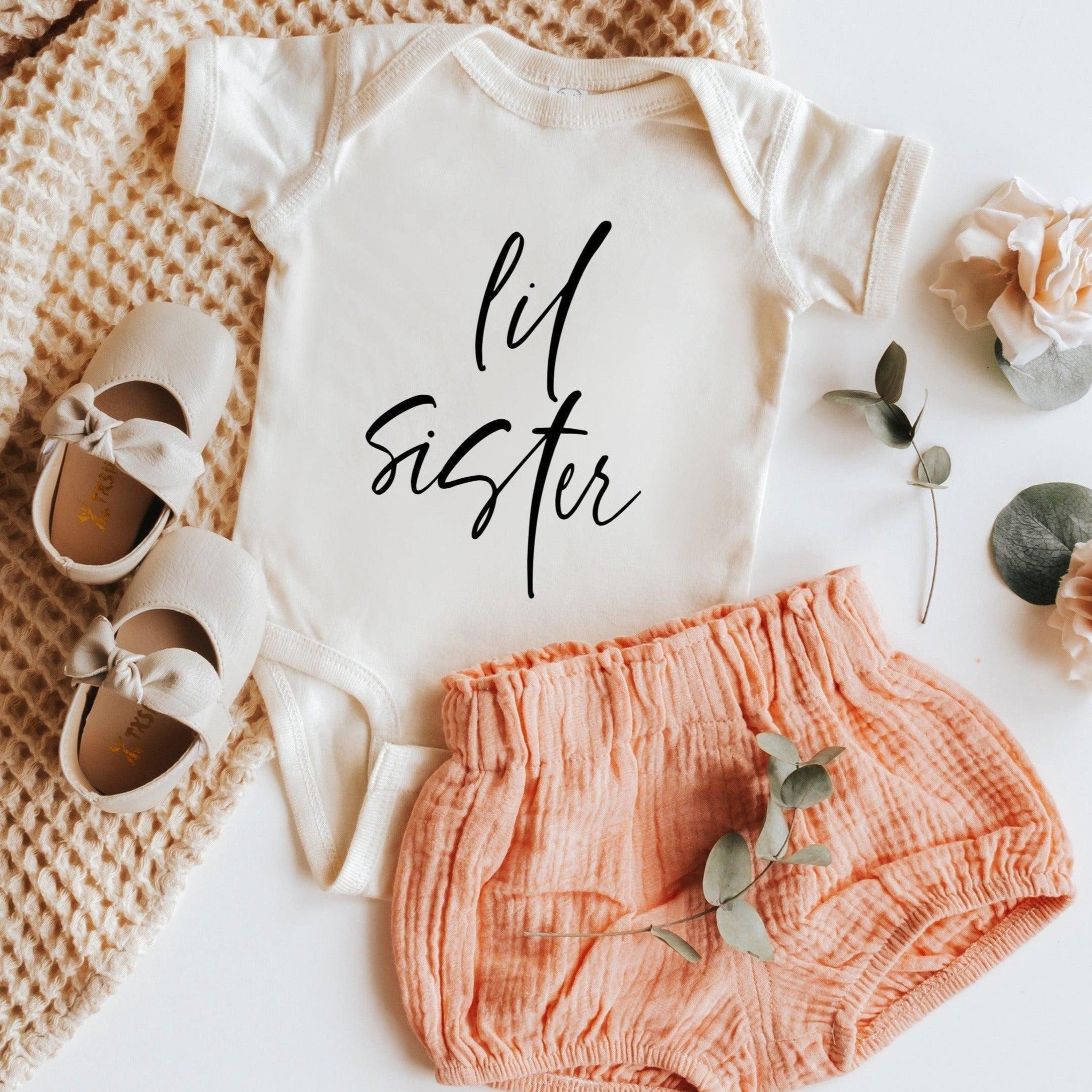 White baby bodysuit with black cursive "lil sister" print