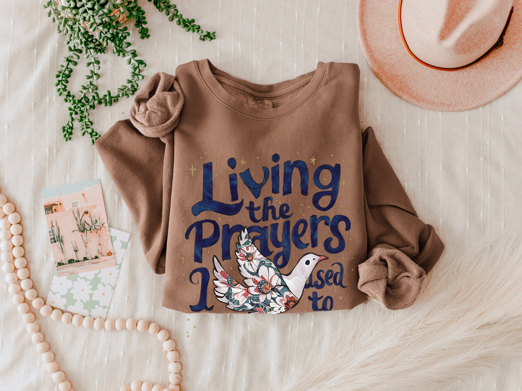 Brown crewneck sweatshirt with "Living the Prayers" floral cross graphic