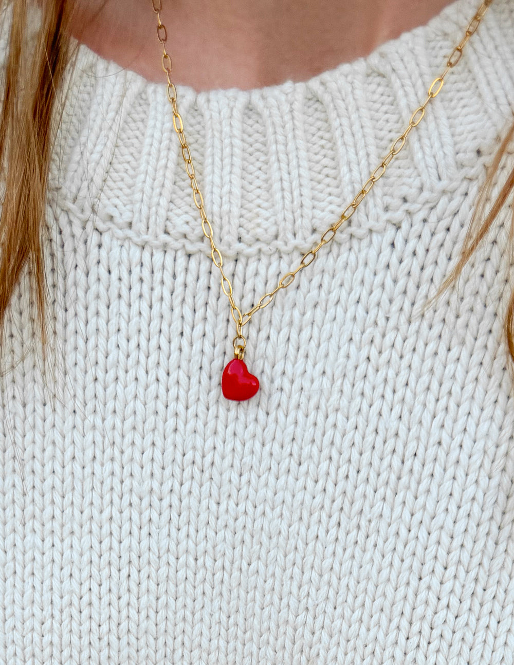 Gold paperclip chain necklace with red heart pendant, close-up on white knit top