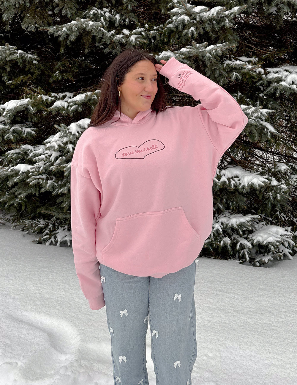 Pink unisex hoodie with hood and heart-shaped “Love Yourself” logo, worn over gray pants.