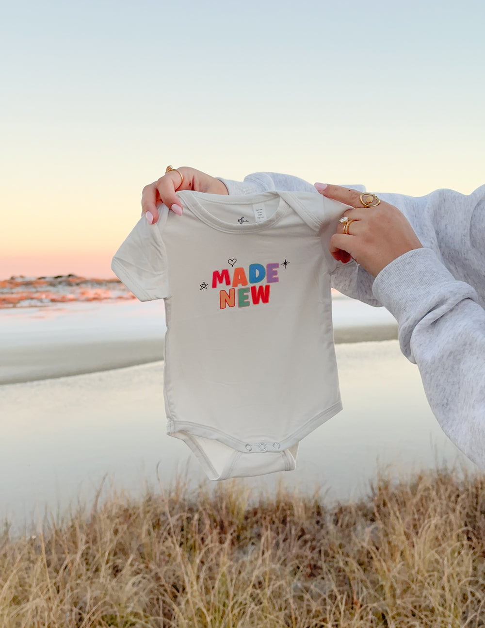 White baby onesie with "MADE NEW" graphic, held by adult hands
