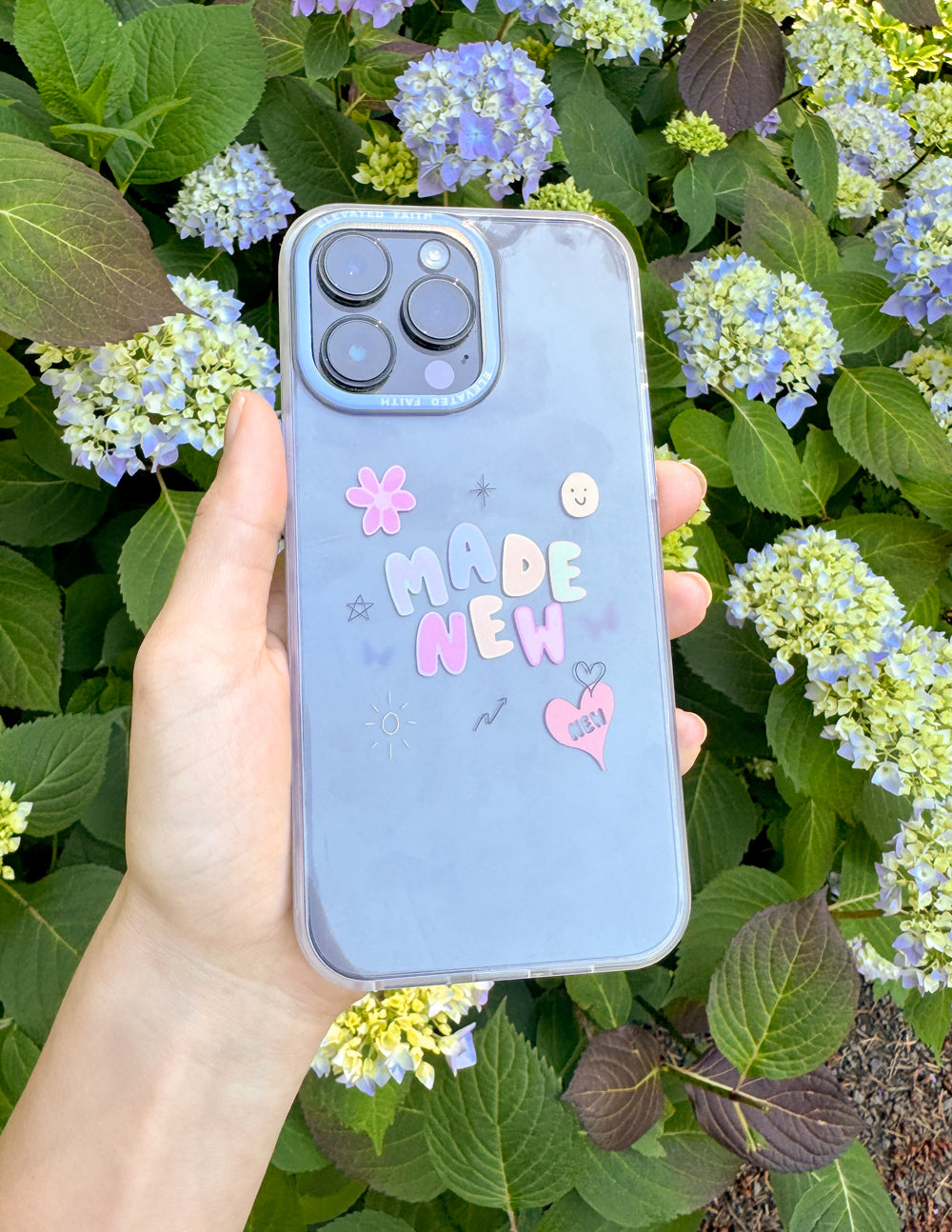 Light blue phone case with 'MADE NEW' graphic, heart and smiley icons, held