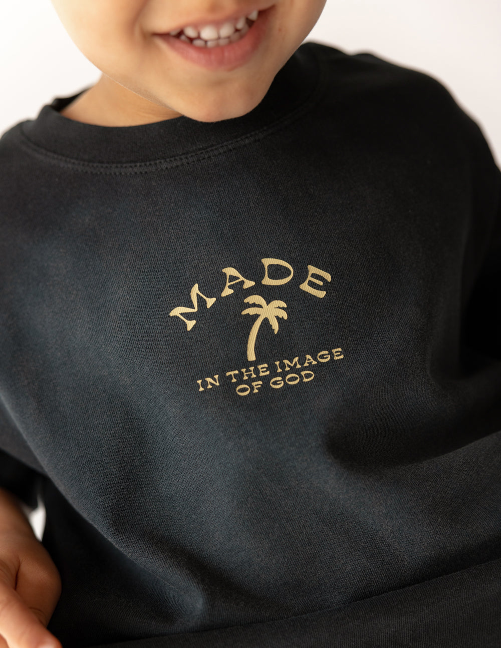 Black kids tee close-up with gold 'MADE' sunburst and 'IN THE IMAGE OF GOD' graphic