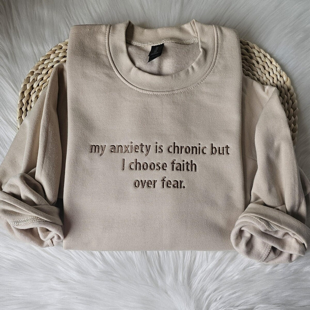 Beige crewneck sweatshirt laid flat with embroidered text: my anxiety is chronic but I choose faith