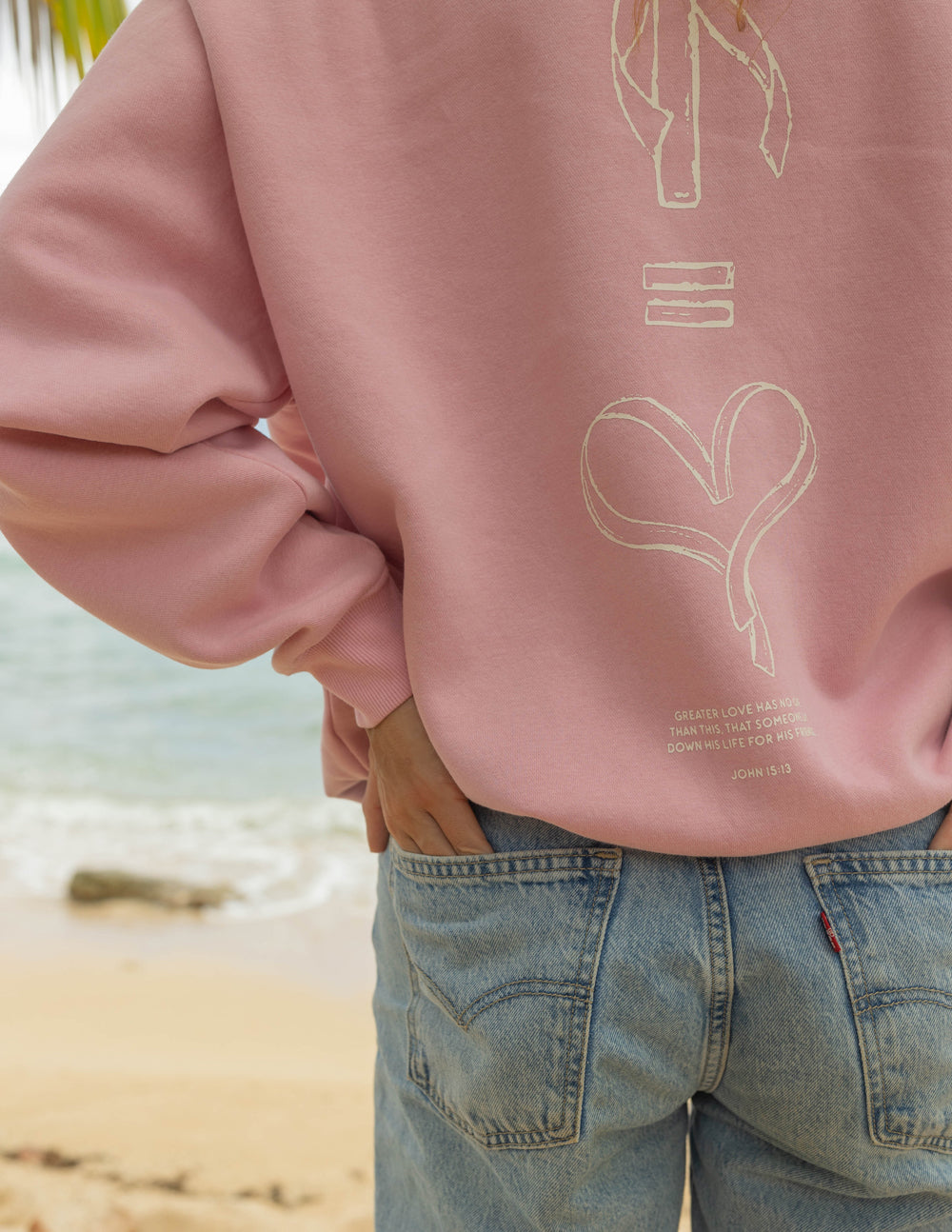Pink sweatshirt back shows bow-and-heart outline graphic with small logo text at lower hem