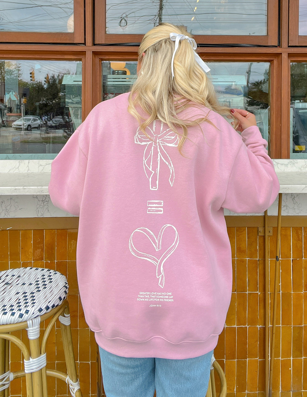 Pink crewneck sweatshirt back view with large bow-and-heart graphic and logo text near hem