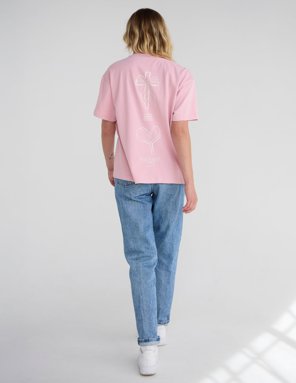 Unisex pink T-shirt back view with small bow and heart line-art graphic.