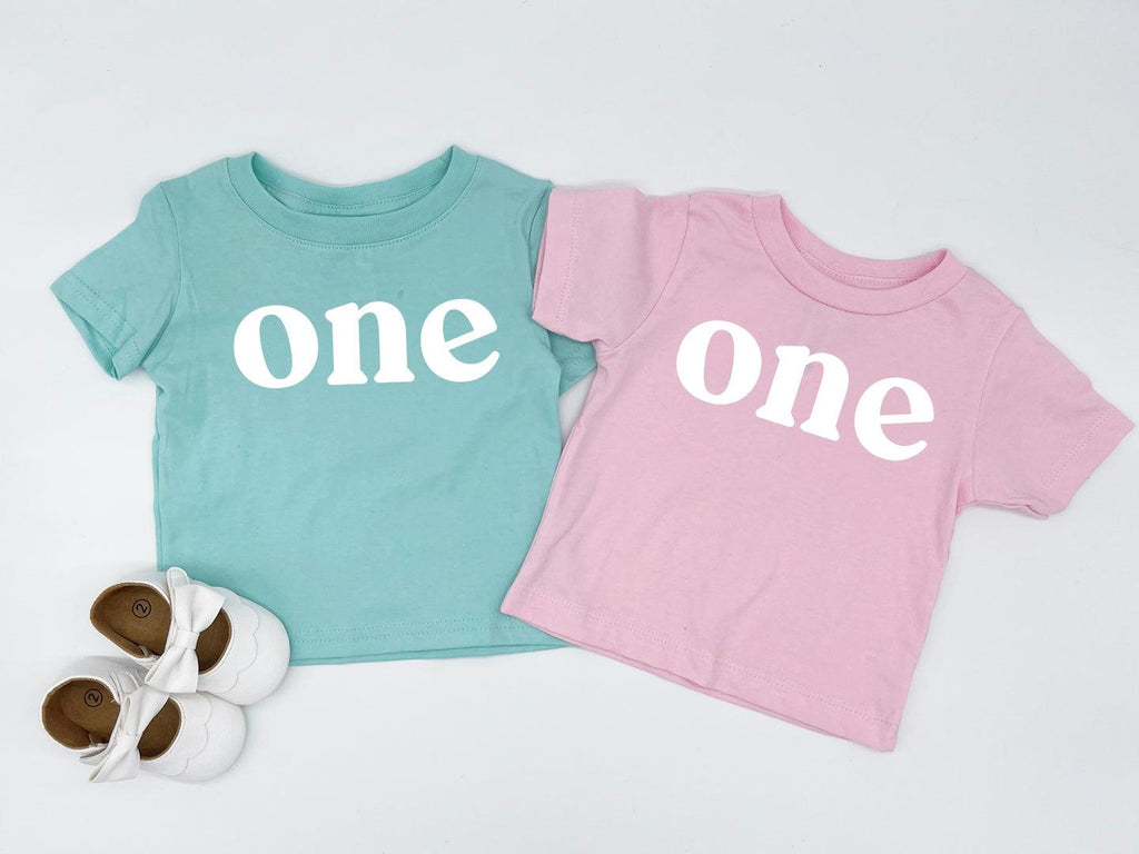 Pair of baby T-shirts: teal “one” and pink “One” serif prints, short sleeves