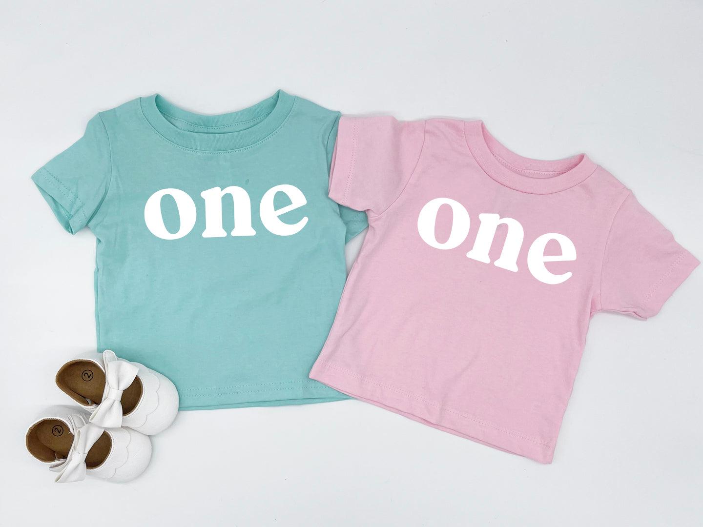 Pair of baby T-shirts: teal “one” and pink “One” serif prints, short sleeves