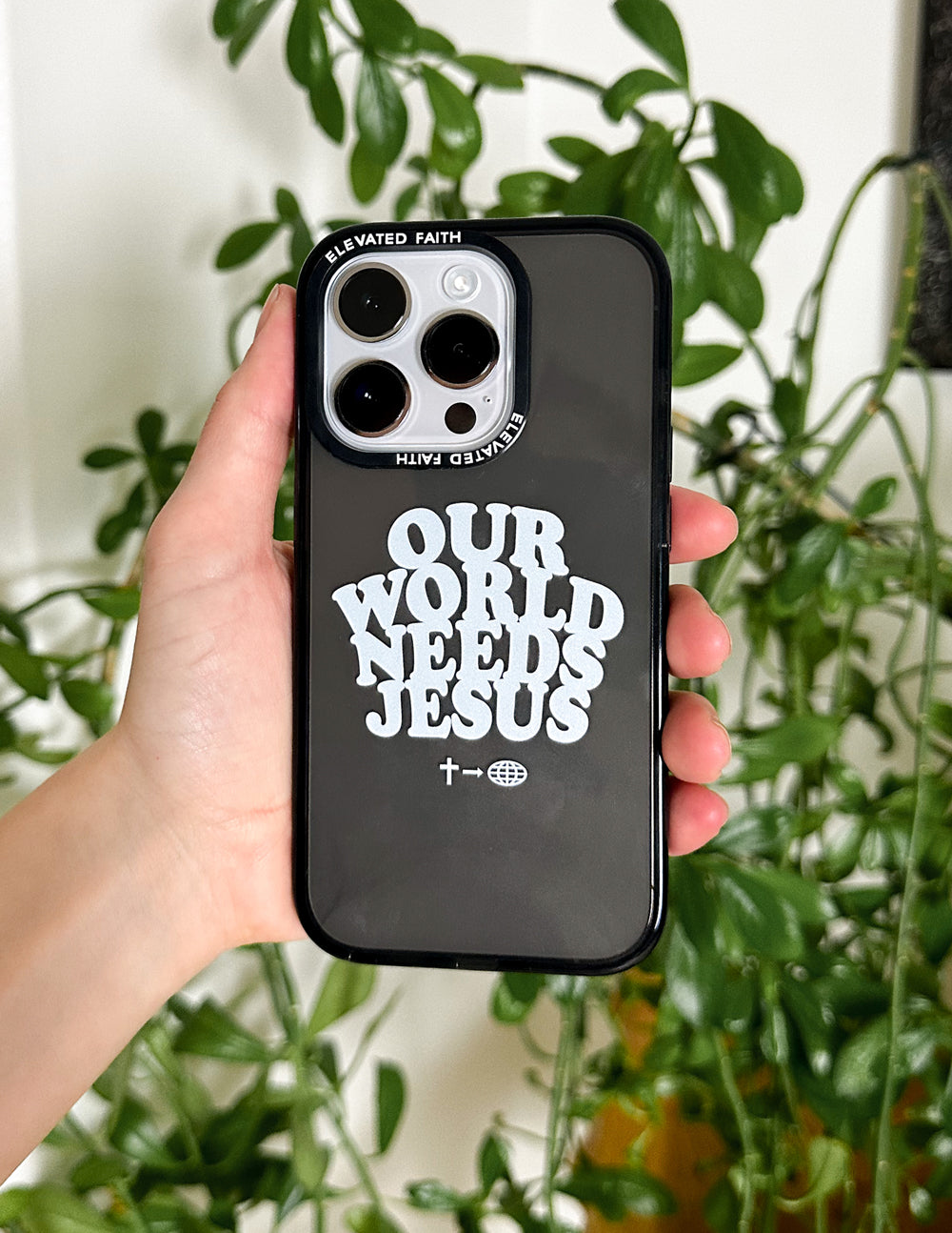 Black phone case with “OUR WORLD NEEDS JESUS” design, held in front of plants