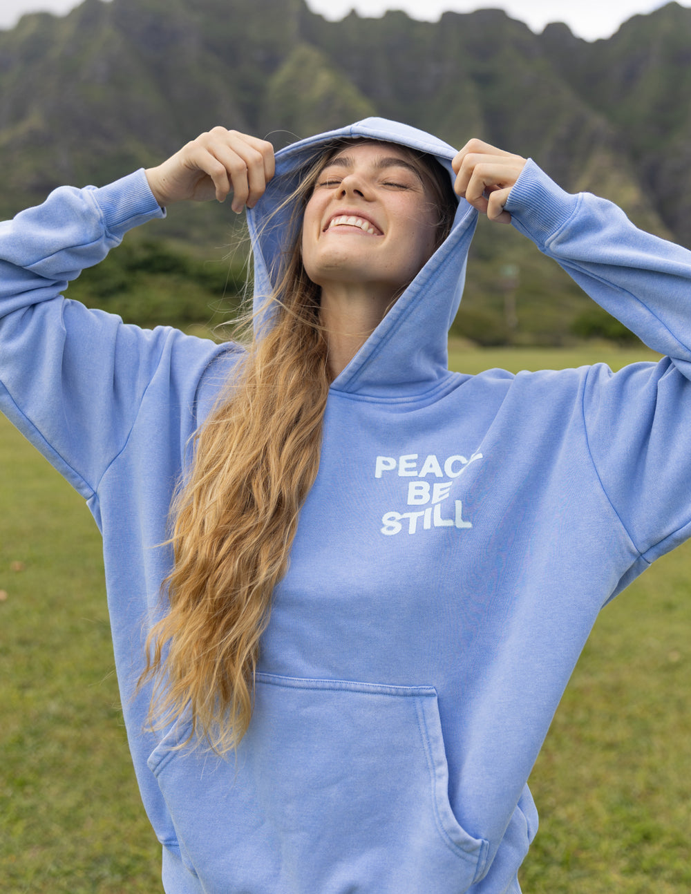 Light blue hoodie with hood up and “PEACE BE STILL” chest graphic, front view