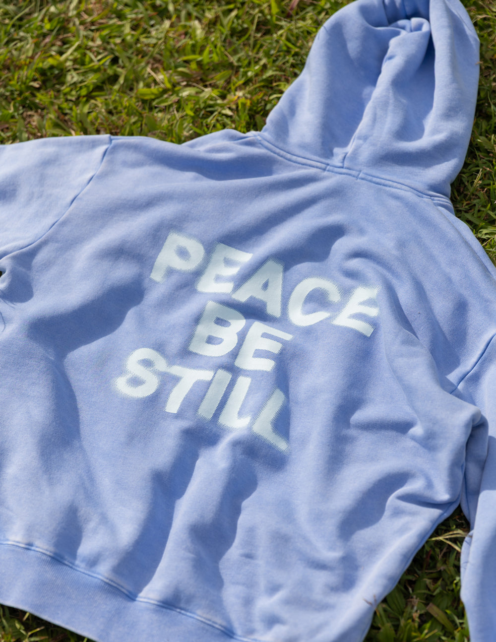 Light blue hoodie sleeve and back graphic reading “PEACE BE STILL”